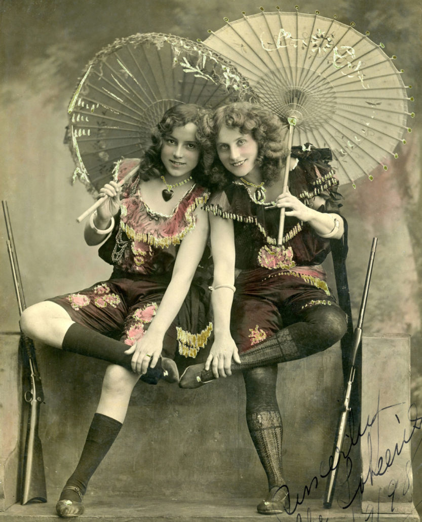 Two girls sitting down holding parasols. There are rifles leaning against the wall