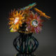 Glass sculpture of multi-colored flowers with faces in the center in a blue wire-like vase