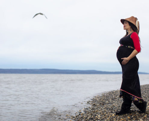Indigenous pregnant woman standing on a rocky shoreline looking out at the ocean