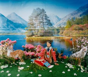Indigenous woman sits in the grass with a red and yellow dress and beaded accessories with a mountain and lake scene behind and cutouts of deer and a wolf around her