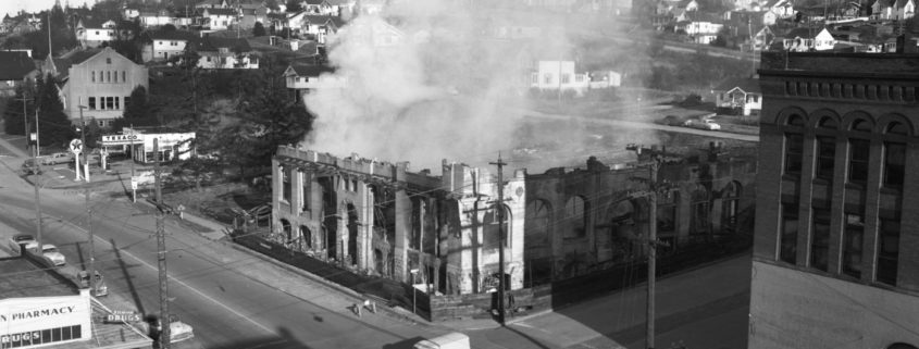 Black and white photo of a building on fire with smoke rising and a hillside with homes around it