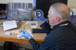 Male identified person with gray hair and a black longsleeved button shirt looks through a viewfinder at a black and white photograph while sitting at a desk with a computer and other photographs