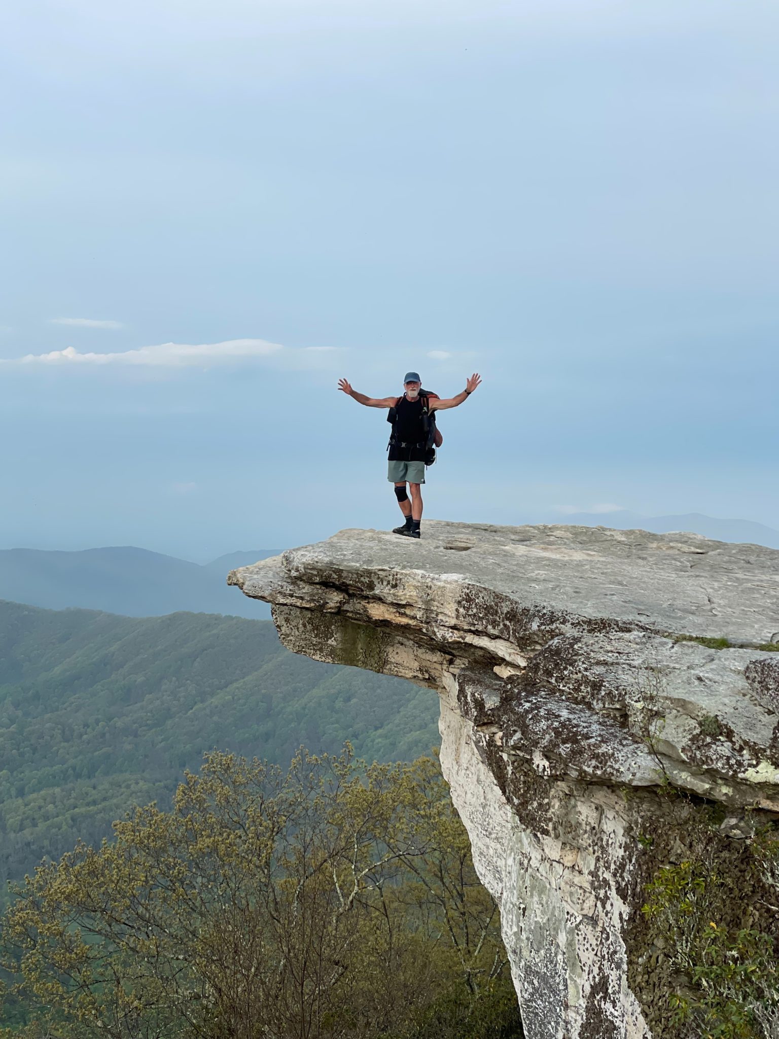 Phot of a man standing on the edge of a rock ledge with his arms raised into the air and a forest and sky in the background