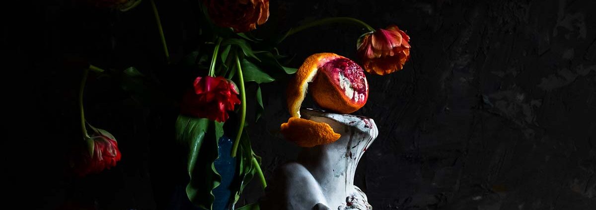 Color still life photograph of a vase with red/orange flowers, a sculpture with a peeled blood orange on top and one squished on the table.