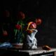 Color still life photograph of a vase with red/orange flowers, a sculpture with a peeled blood orange on top and one squished on the table.